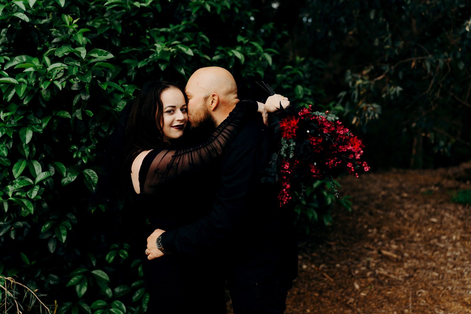 A romantic couple embraces on a wooded path surrounded by lush greenery and natural light.