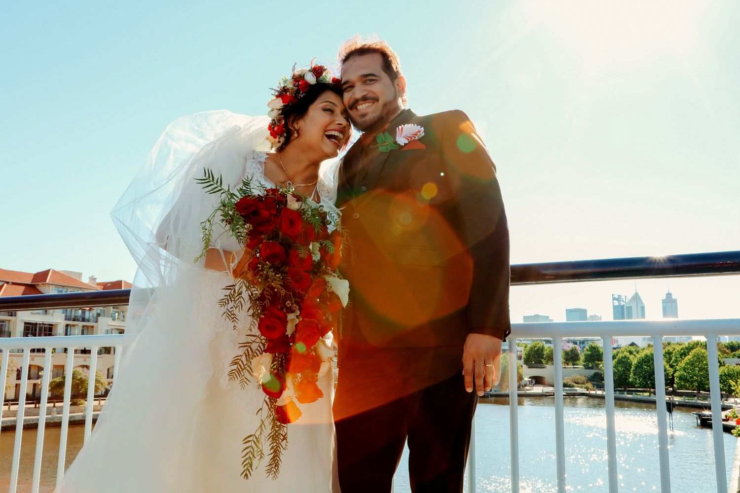 Wedding couple embraces on a balcony with vibrant red floral arrangements against a blue sky backdrop.