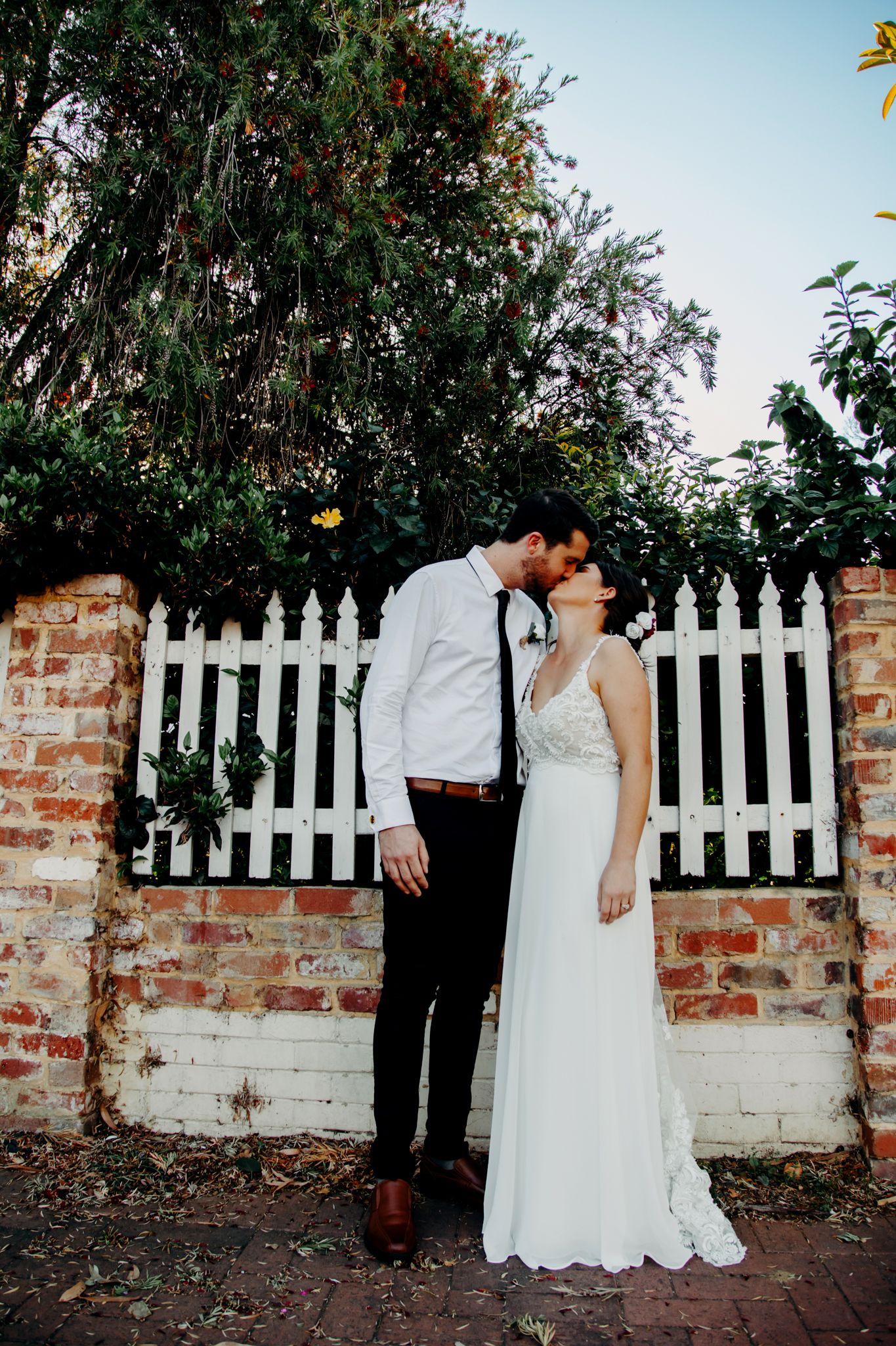 Romantic wedding portrait against white picket fence and brick wall at dusk.