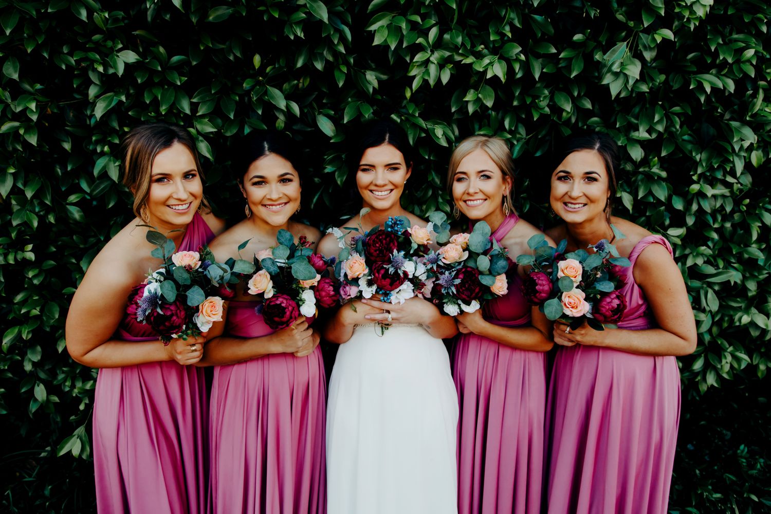 Bridal party in pink dresses holding colorful bouquets against ivy wall backdrop.