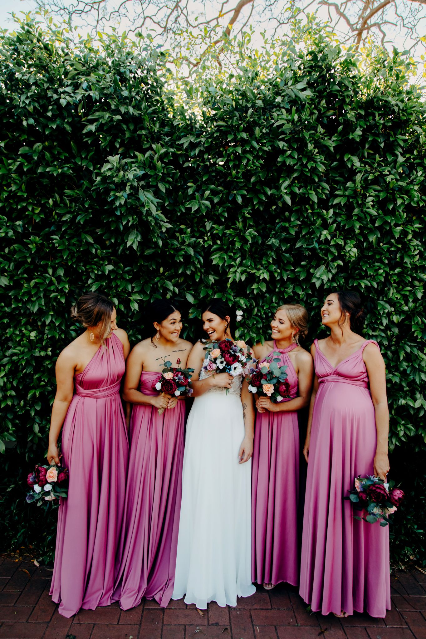 Bridal party in pink maxi dresses posing against lush green hedge wall.