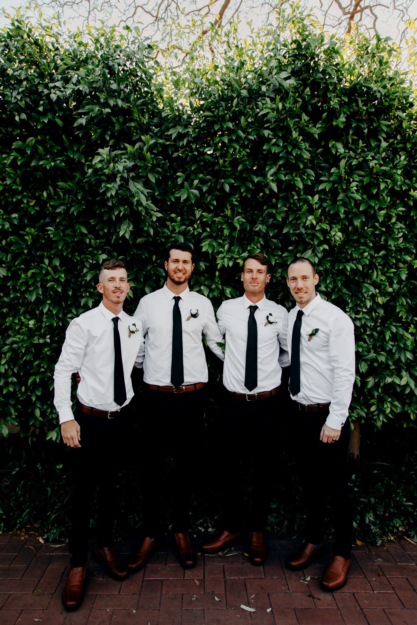 Groomsmen in matching white shirts and black ties pose against lush green hedge wall.