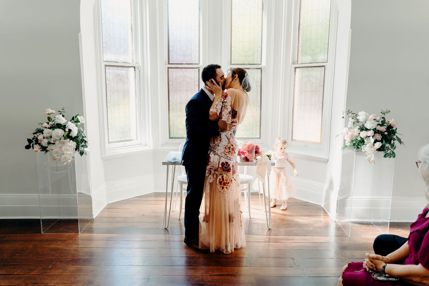 A couple shares an intimate dance in a sunlit room with white decor and floral arrangements.