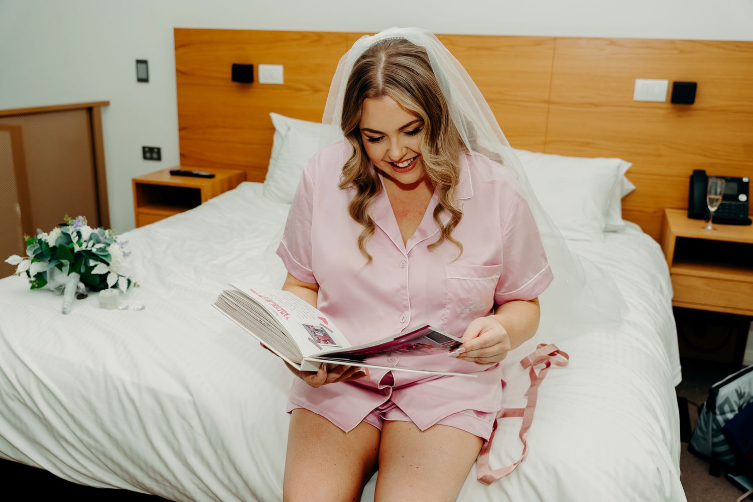A person in pink sitting on a bed reads while wearing a wedding veil.