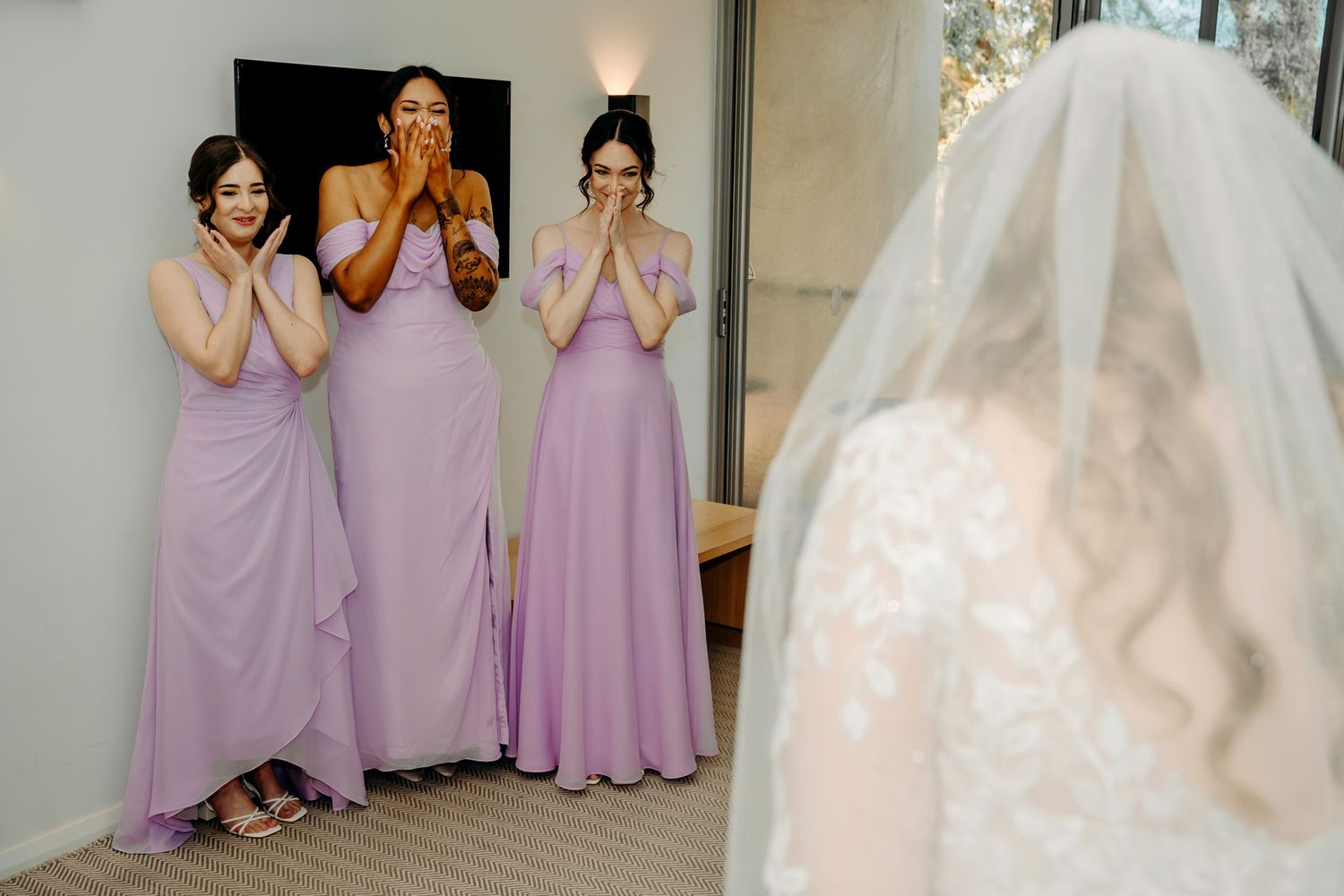 Bridesmaids in lavender dresses react with joy to first look at the bride.