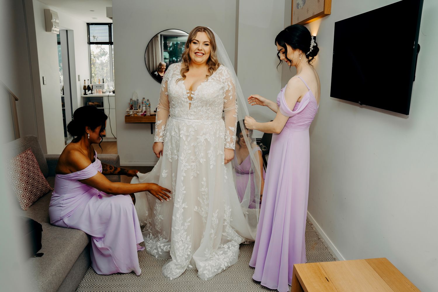 Bridesmaids helping with final wedding dress adjustments in a bright room.