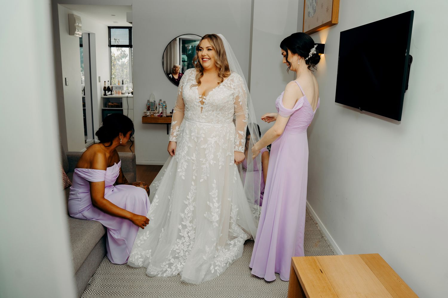 Wedding dress fitting scene in bright modern room with bridesmaids in pastel dresses.
