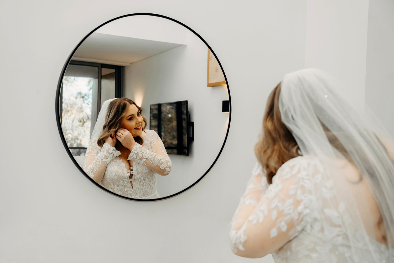 A bride checking her reflection in a round mirror while putting on earrings.