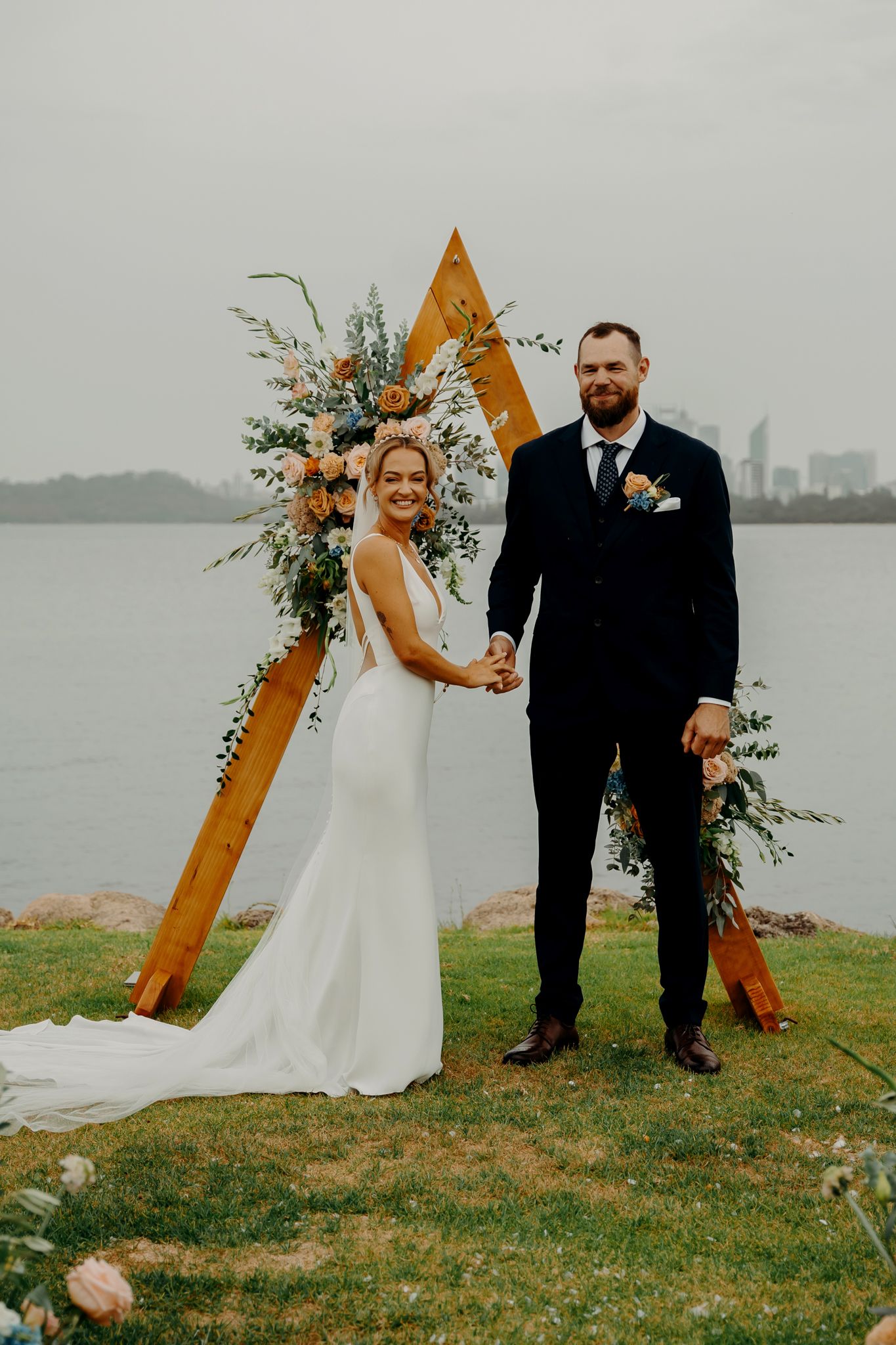 A modern waterfront wedding featuring a geometric wooden arch decorated with fresh florals.