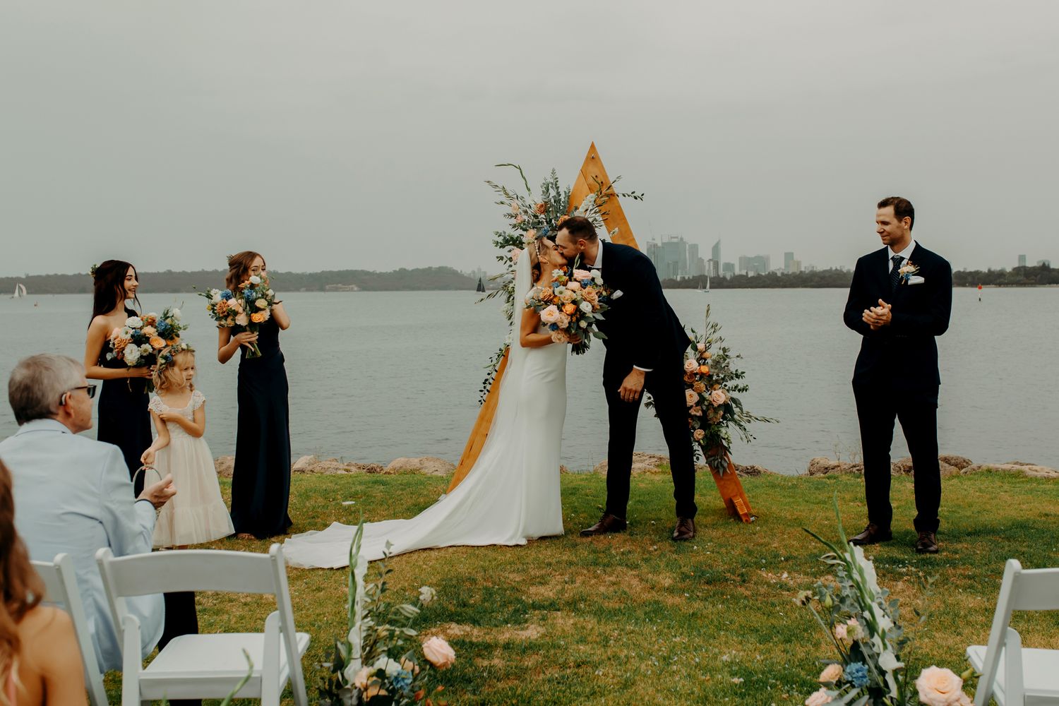 Outdoor wedding ceremony setup with wooden triangle arch and white chairs by the water.