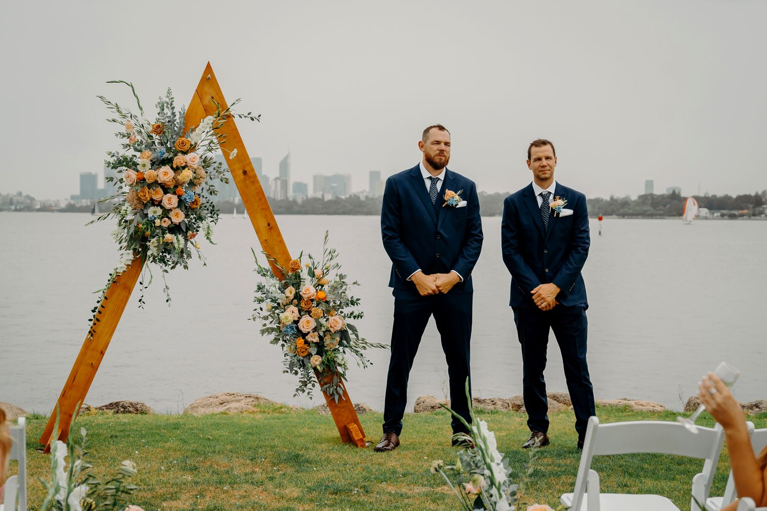 Two groomsmen stand beneath a wooden triangle arch decorated with flowers at a waterfront wedding ceremony.
