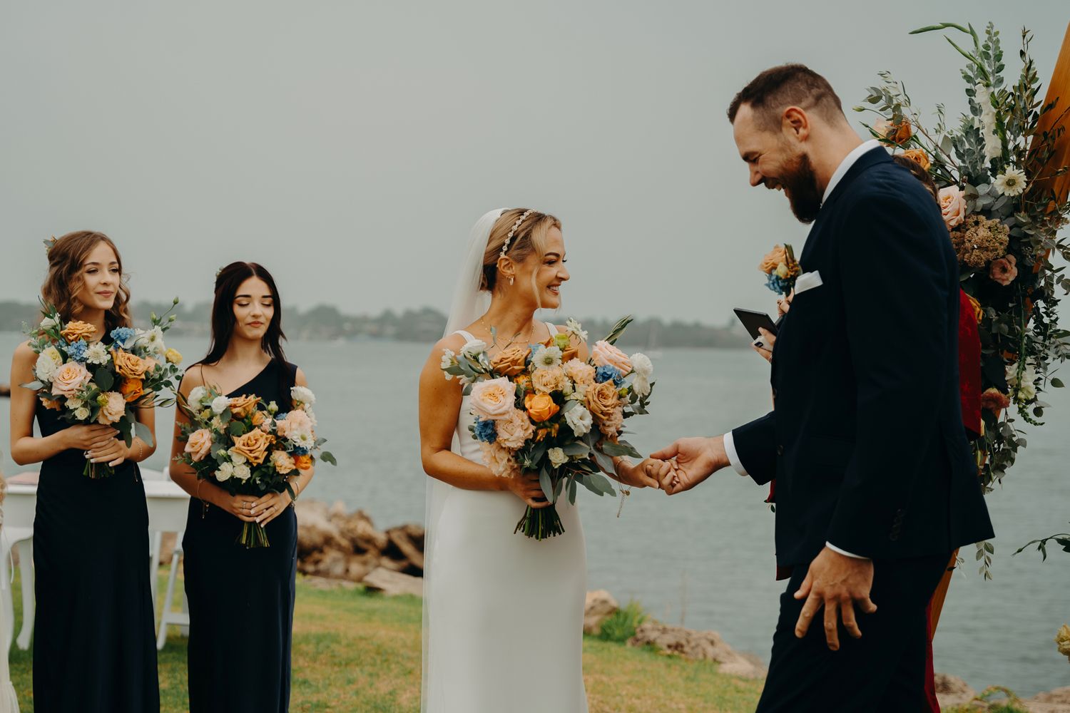 An intimate waterfront wedding ceremony with bridesmaids in navy dresses and floral arrangements.