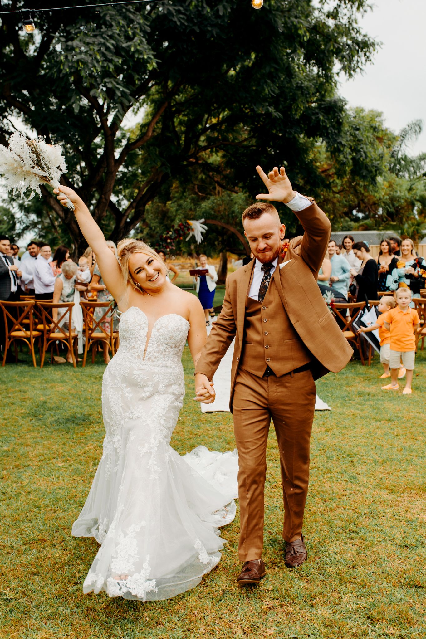 Joyful newlyweds celebrate at an outdoor garden wedding reception with raised arms and excited expressions.