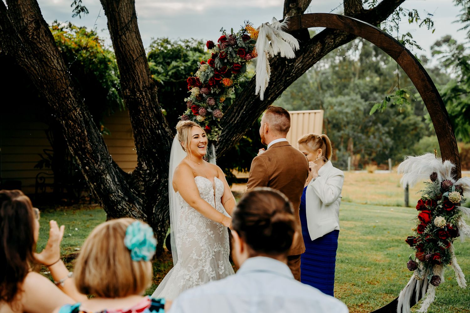 A lovely outdoor wedding ceremony taking place under a rustic wooden arch adorned with pink and white floral arrangements.