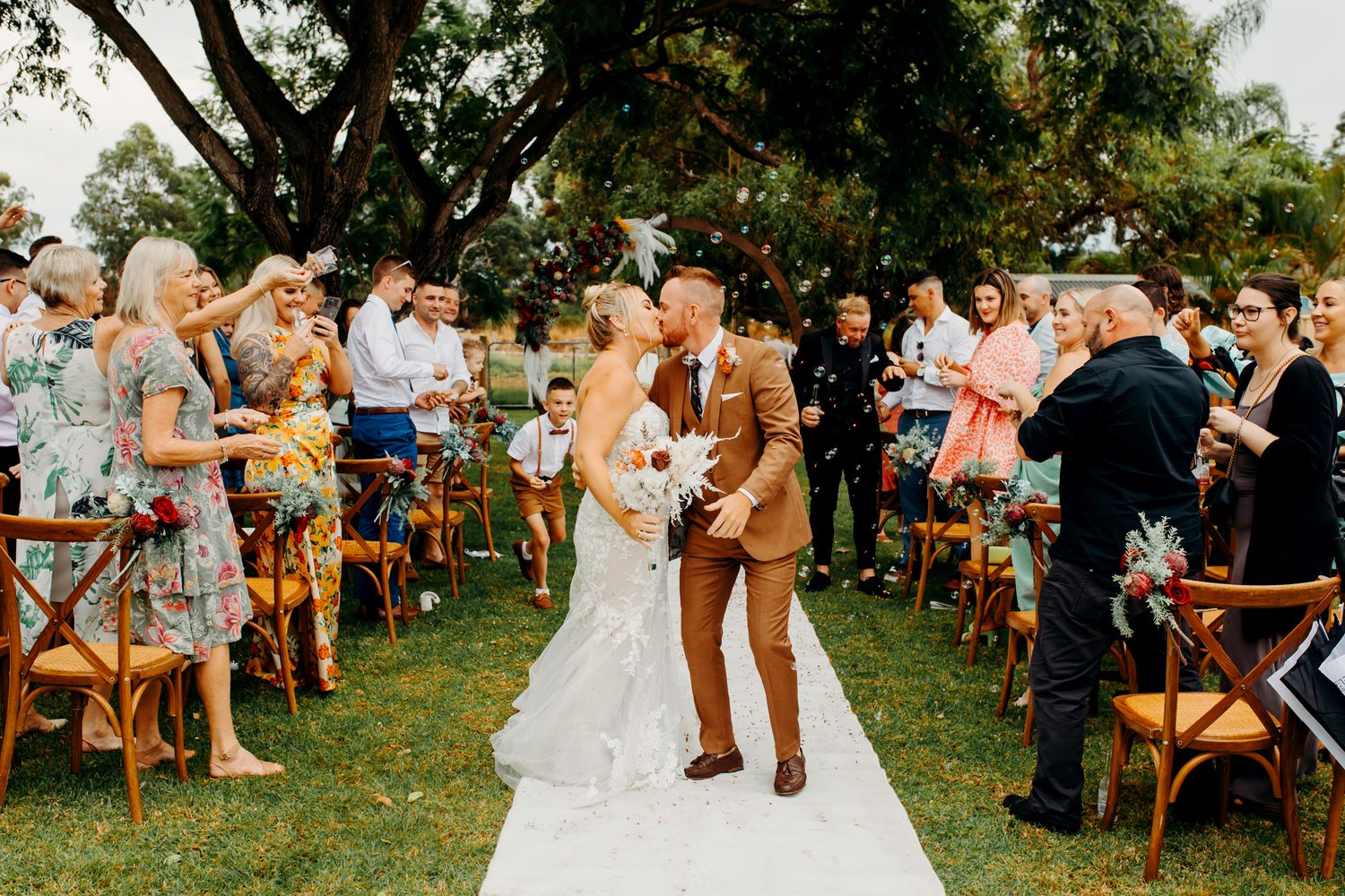 A joyful outdoor wedding ceremony with wooden chairs and a white aisle runner surrounded by green trees and guests.