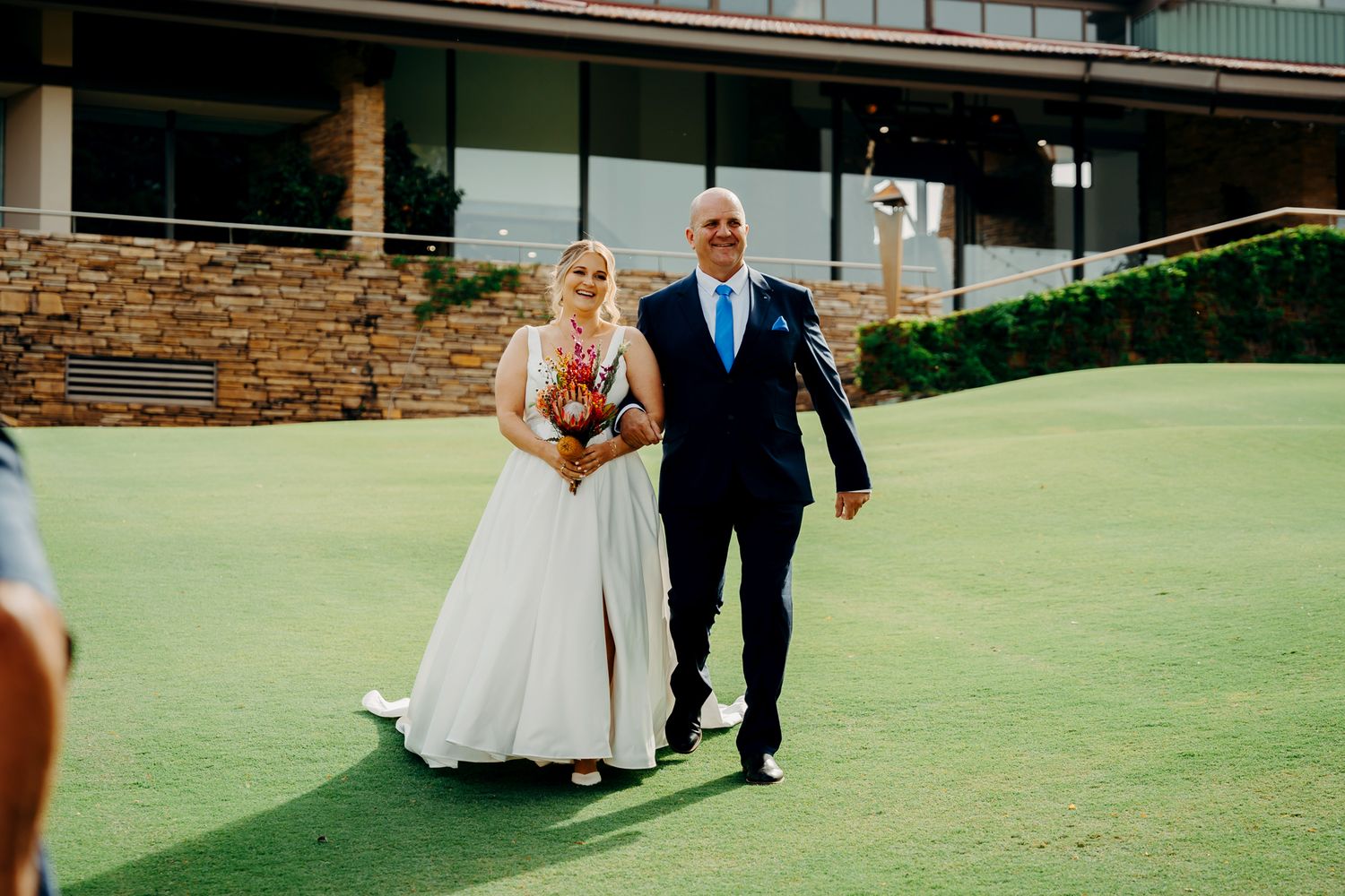 A wedding couple walks together on a lush green golf course with stone building in background.