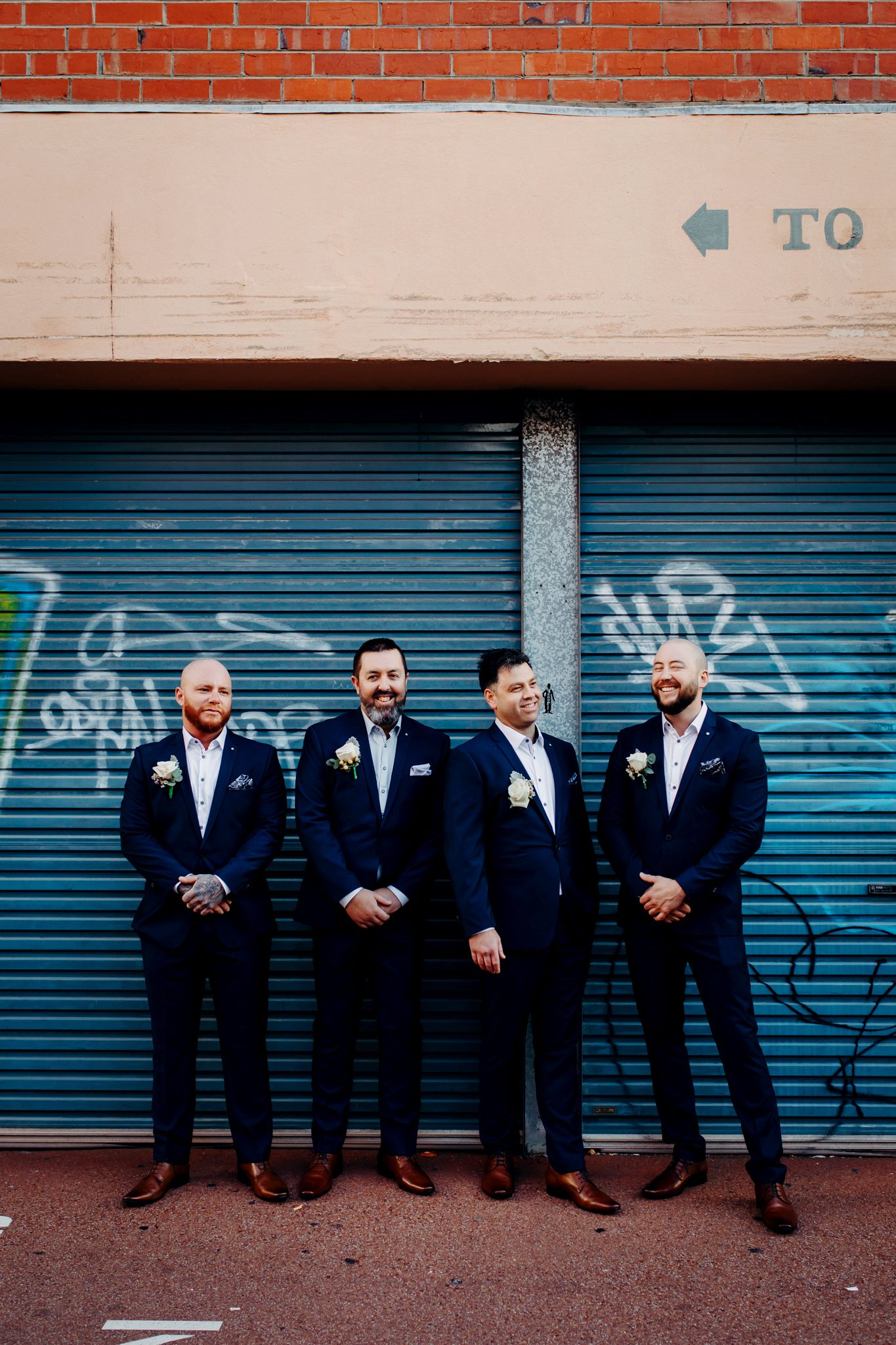 Groomsmen in navy suits posing against industrial blue garage door and brick wall.