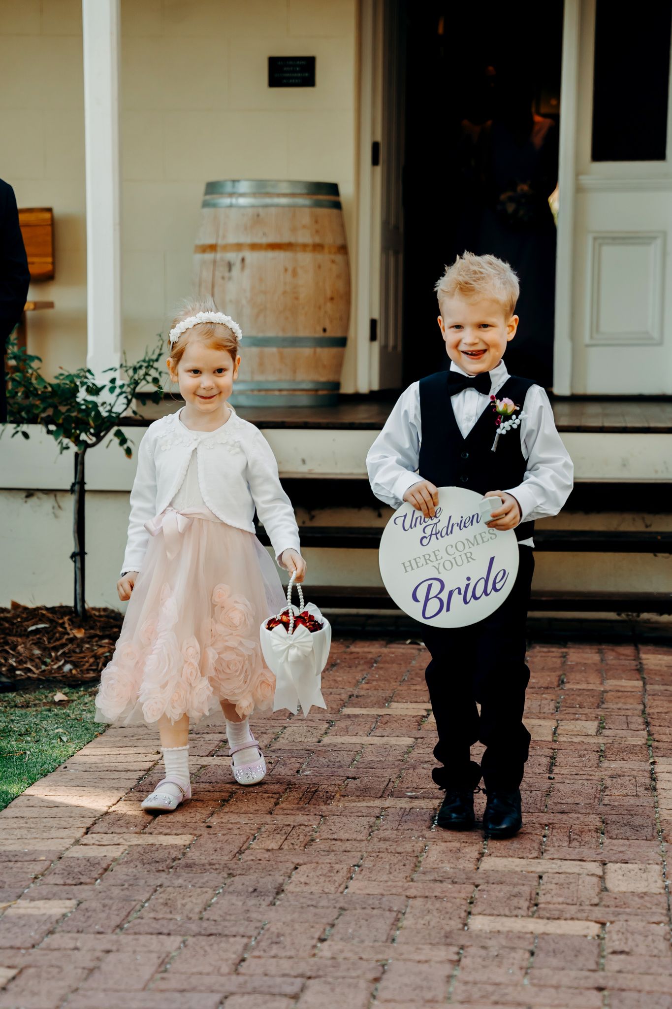 Two young children in formal attire on brick path with wedding signage and basket.