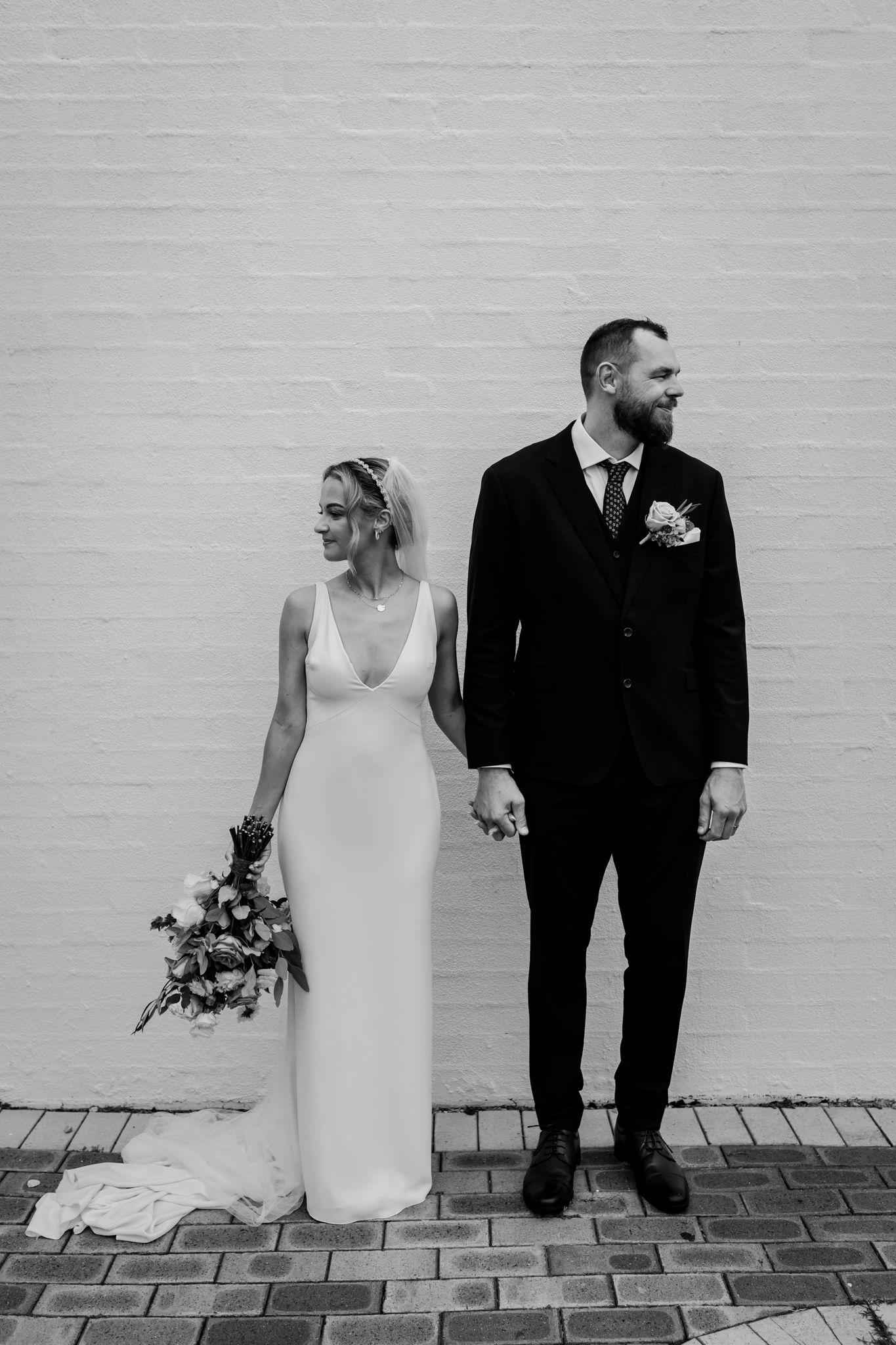 Black and white wedding portrait of couple standing against white brick wall with bride holding floral bouquet.