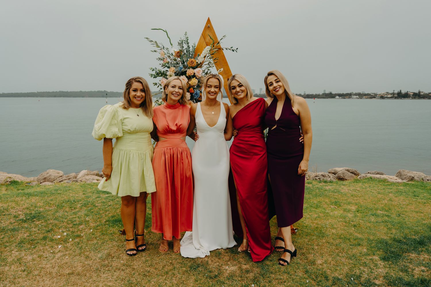 Wedding party poses by triangular arch with bridesmaids in colorful dresses at waterfront venue.