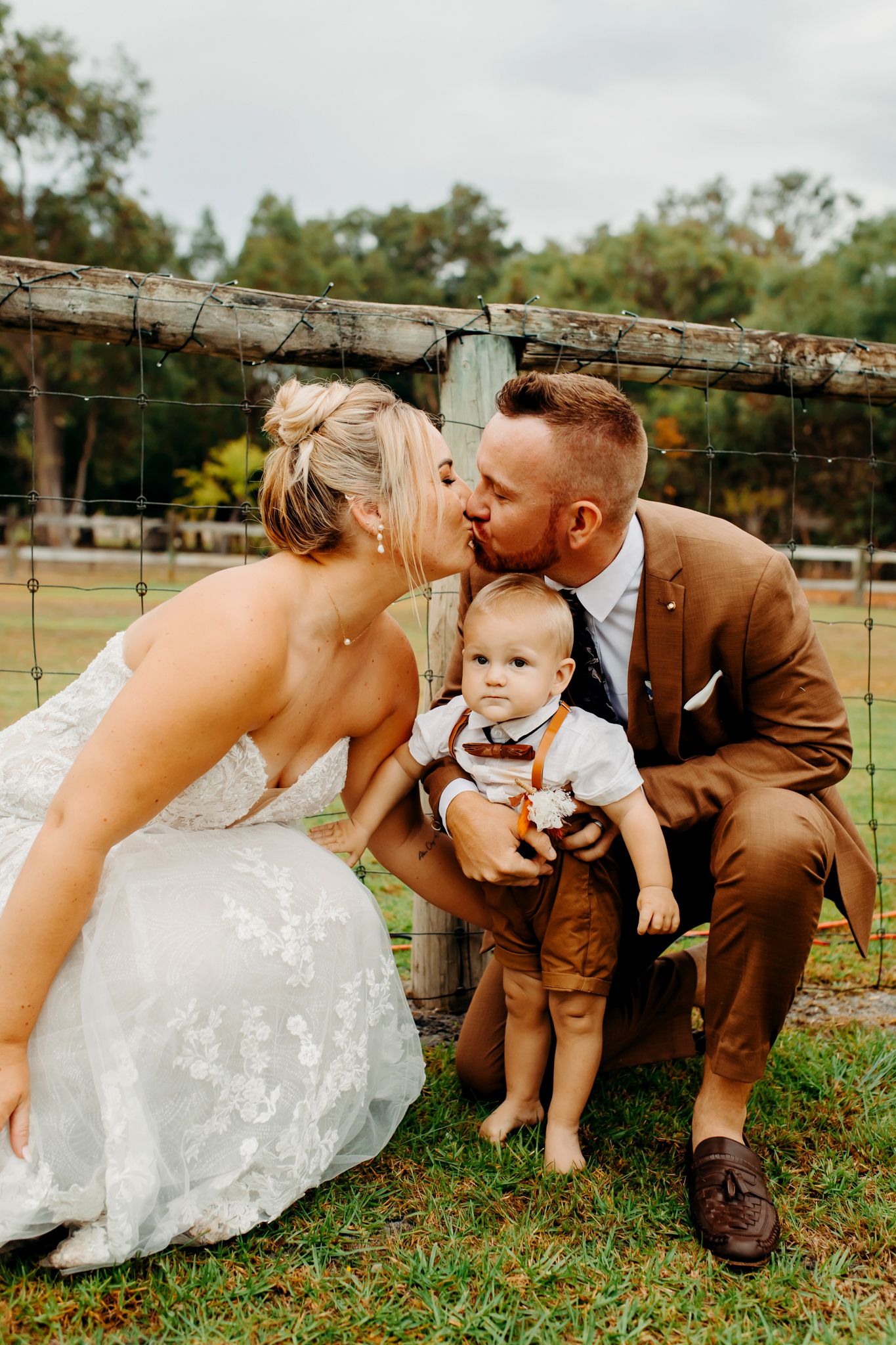Wedding couple shares a kiss while holding small baby in brown outfit outdoors by rustic wooden fence.