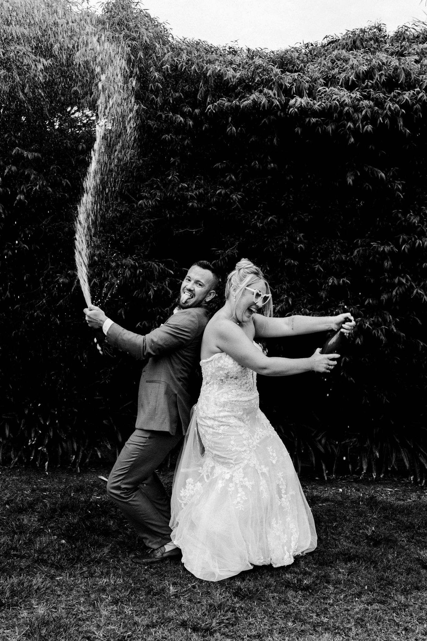 A bride and groom celebrate with champagne sprays while standing back-to-back in a garden setting.