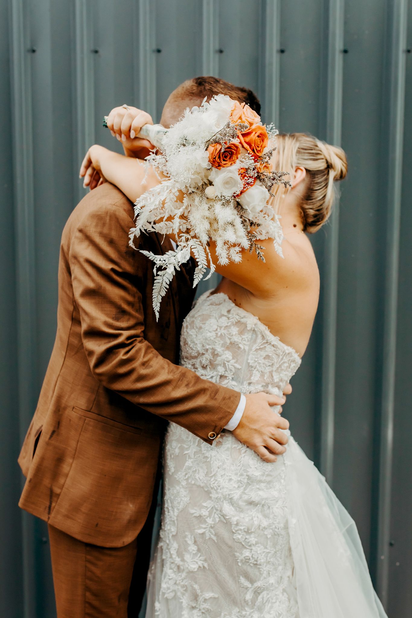 A wedding couple embraces while holding a bouquet with white and orange flowers against a teal metal wall.