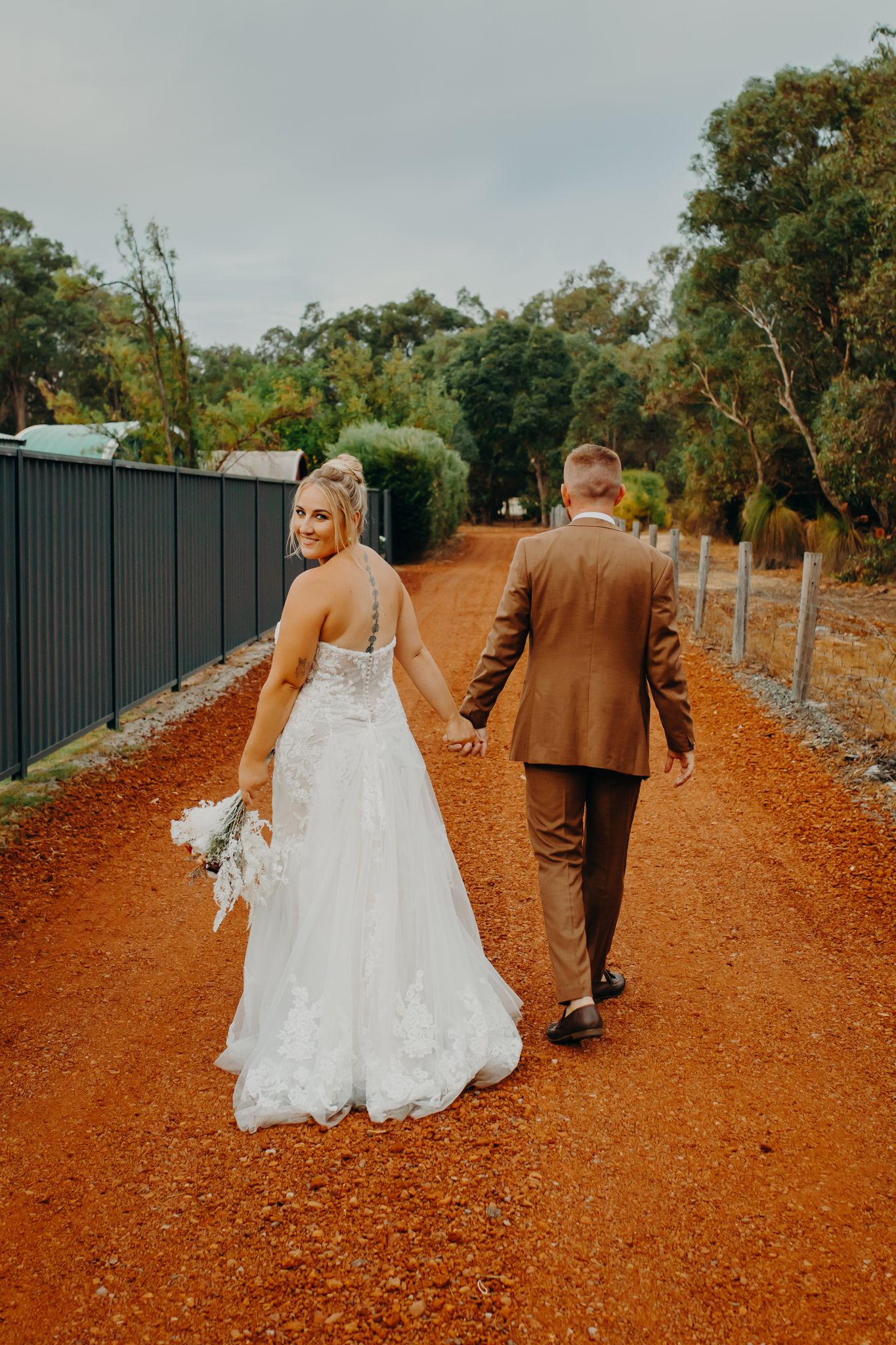 Newlyweds walk down a rustic red dirt path lined with trees while holding hands on their wedding day.