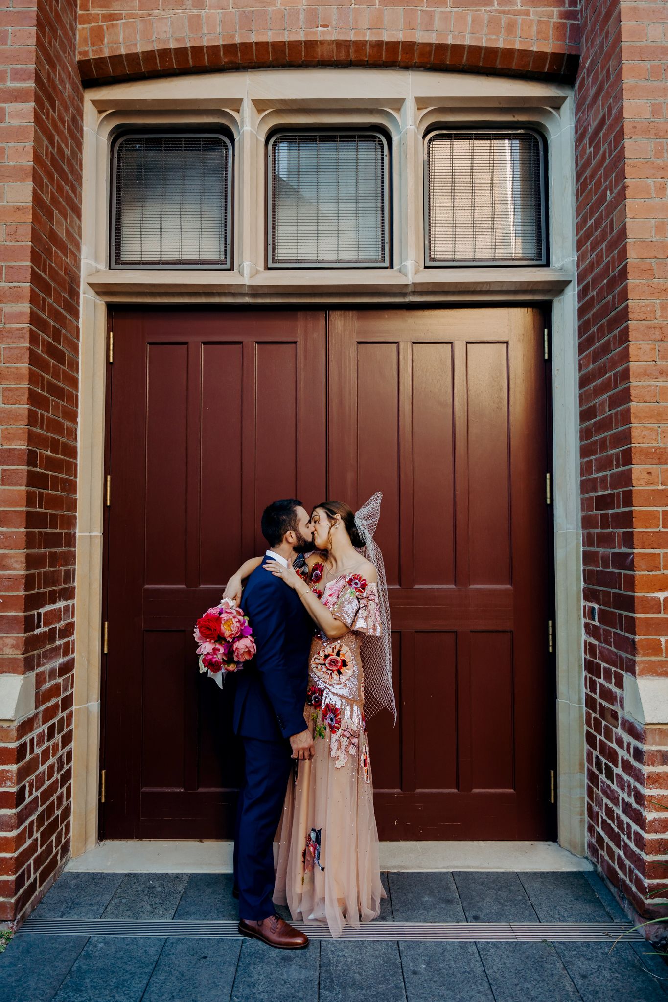 A couple poses romantically in front of wooden doors in a brick archway.