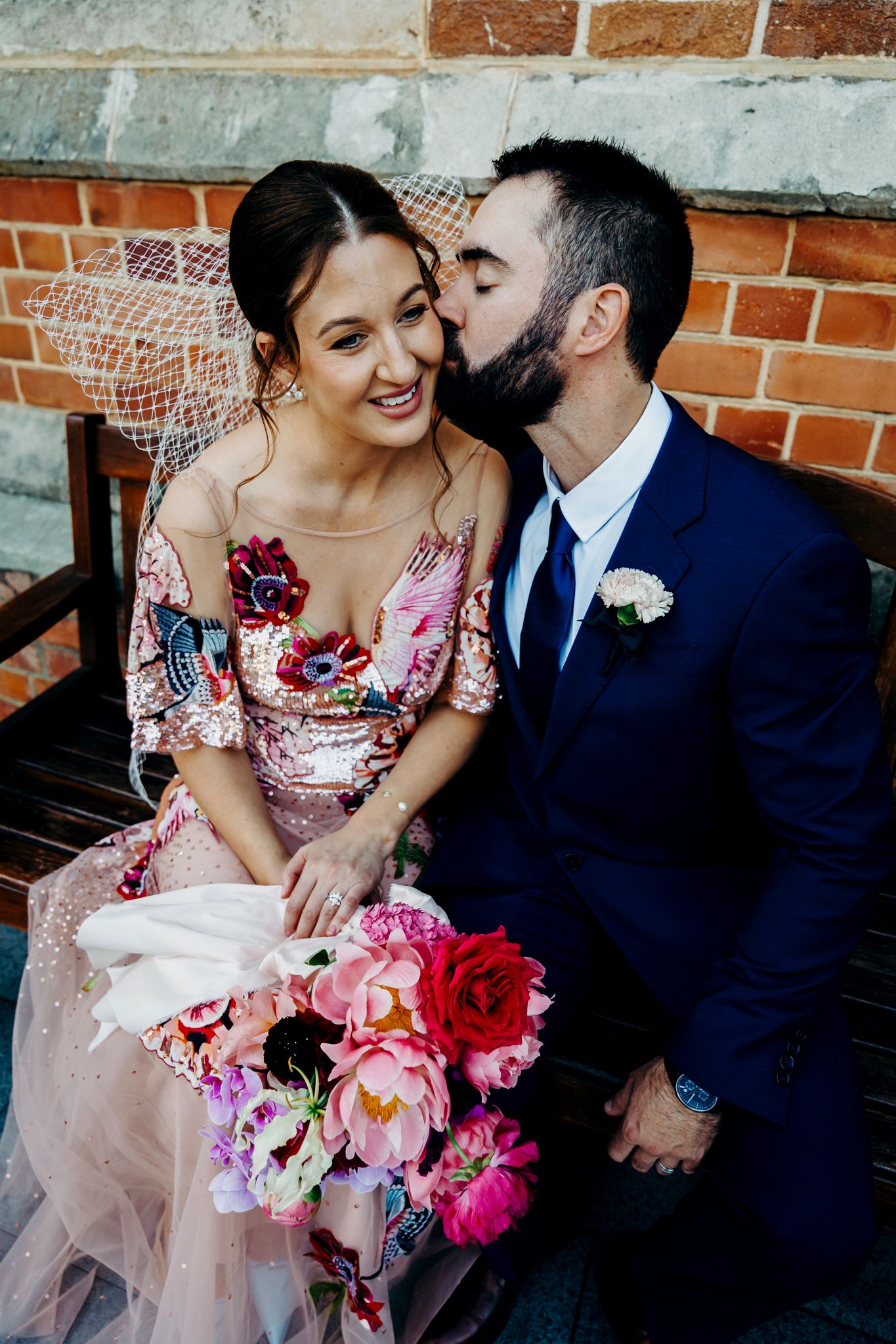 A couple shares an intimate moment on a bench with a vibrant pink floral bouquet.