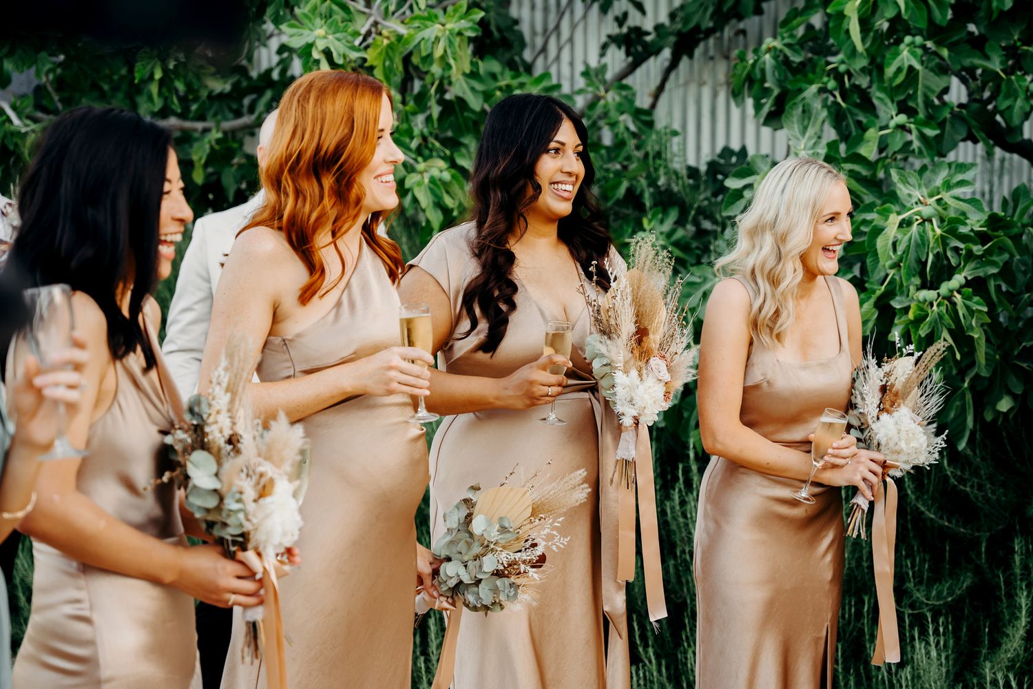 Bridesmaids wearing elegant champagne satin dresses hold bouquets while standing in line at an outdoor wedding.