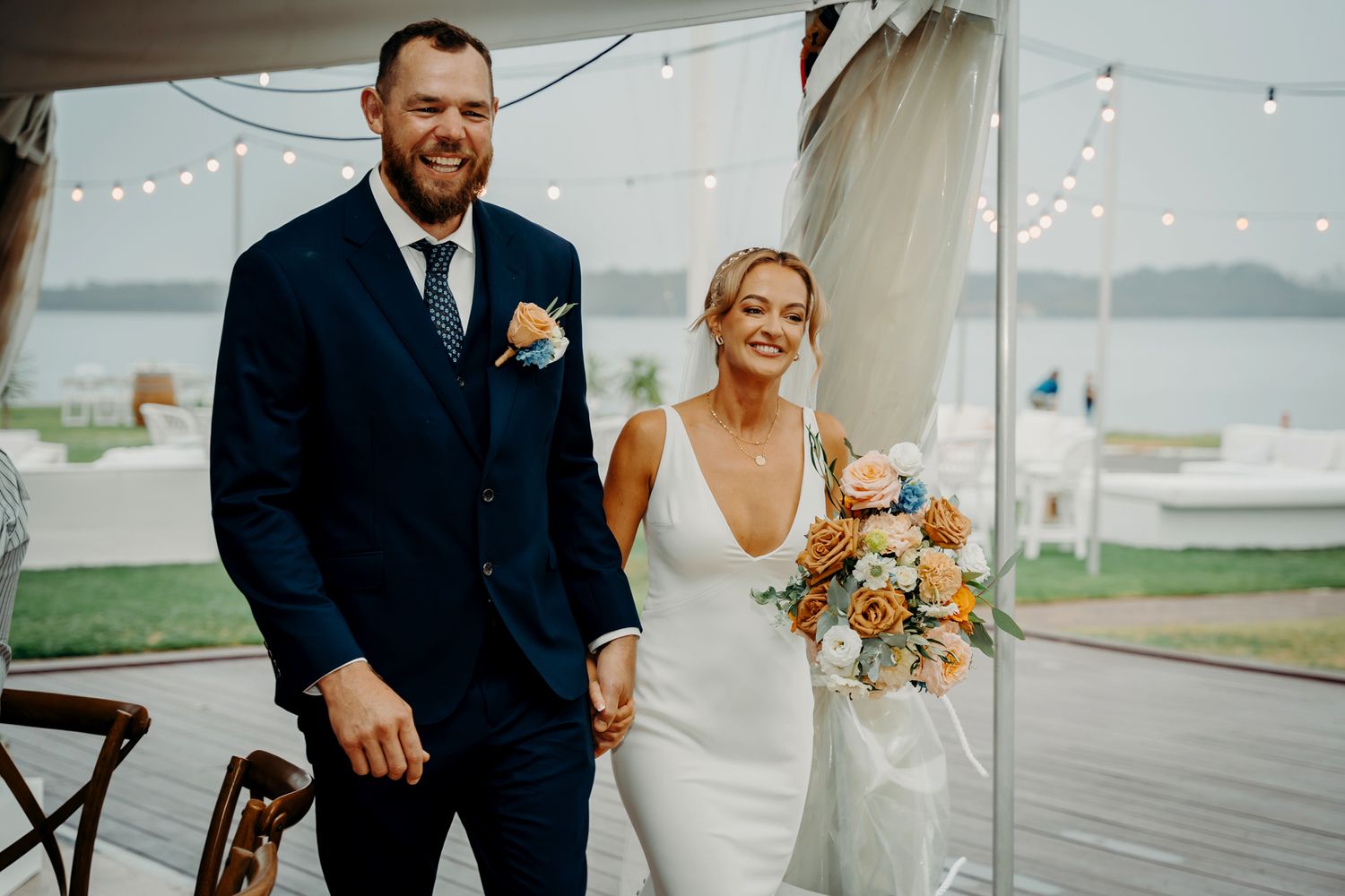 Wedding couple poses on waterfront deck with string lights and peach floral bouquet.