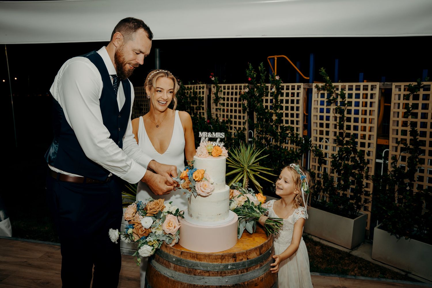 Couple cuts wedding cake with flower girl at evening reception.