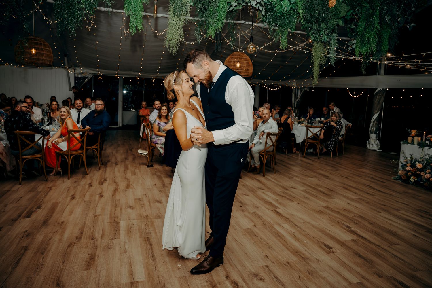 First dance at wedding reception with greenery decor and wooden dance floor.