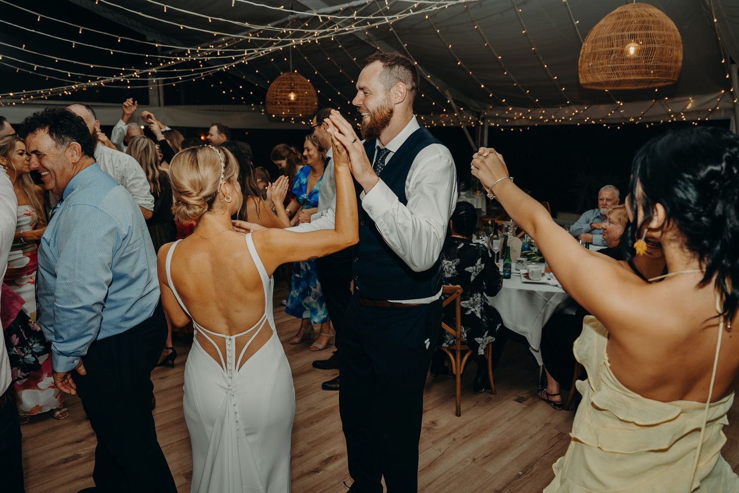 Wedding guests dance and celebrate on a wooden dance floor under string lights.