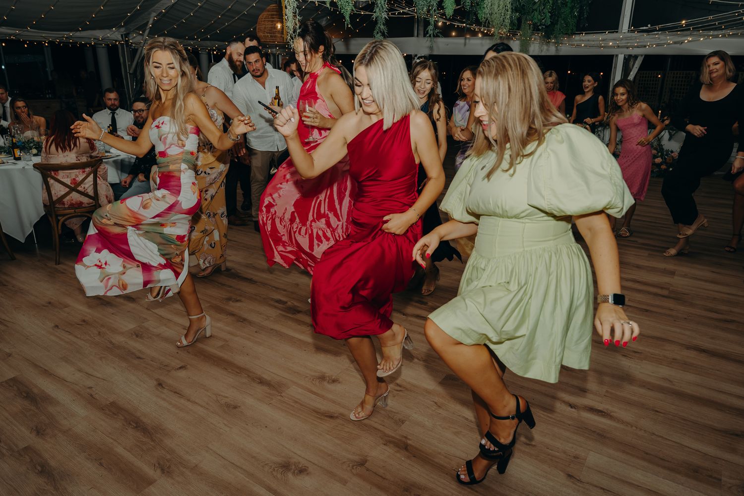 Wedding guests dancing and twirling in colorful dresses on a wooden dance floor.