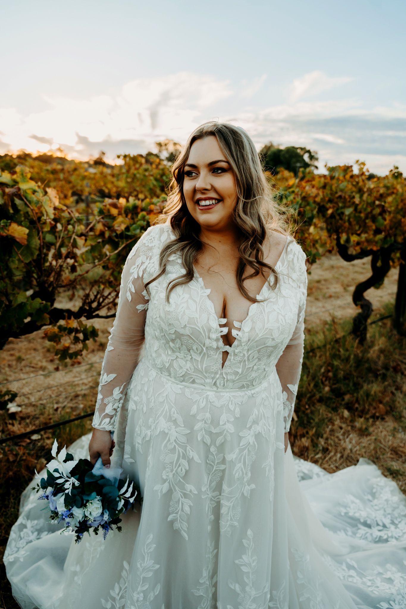 A bride in a lace wedding gown stands among autumn vineyard vines.