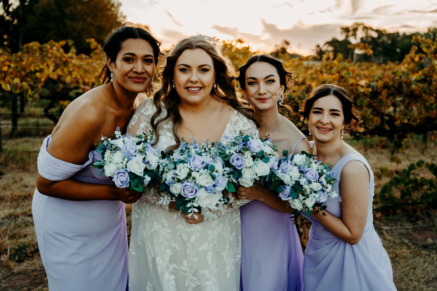 Wedding party with blue bouquets poses together in a vineyard at sunset.