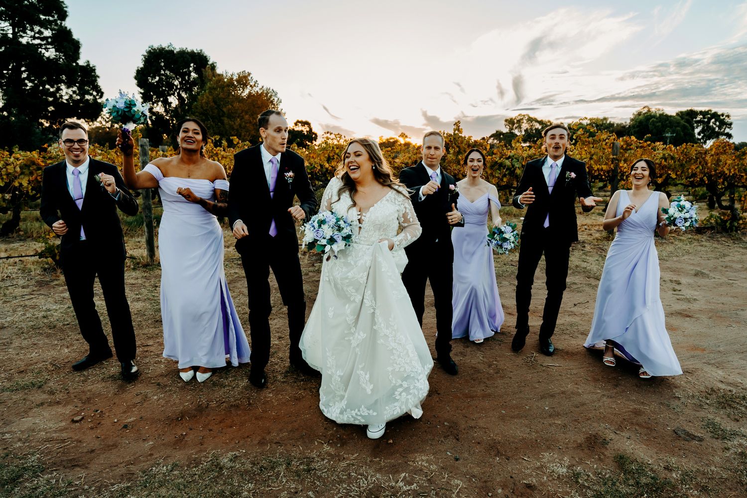 Wedding party poses together in vineyard at sunset with bridesmaids in lavender dresses and groomsmen in dark suits.