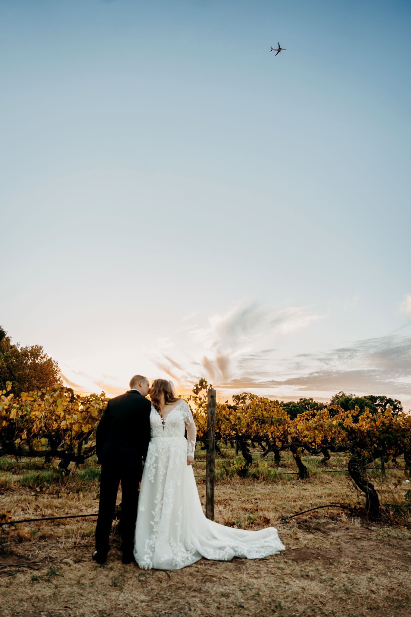 Couple shares a moment in vineyard with small airplane visible in bright blue sky.