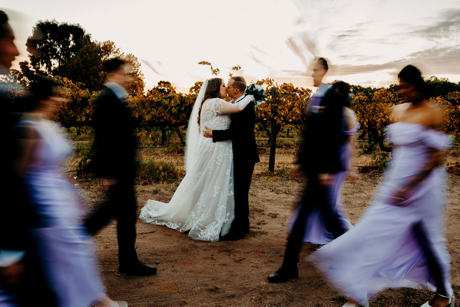 Motion-blur wedding party circles around couple in vineyard at dusk.