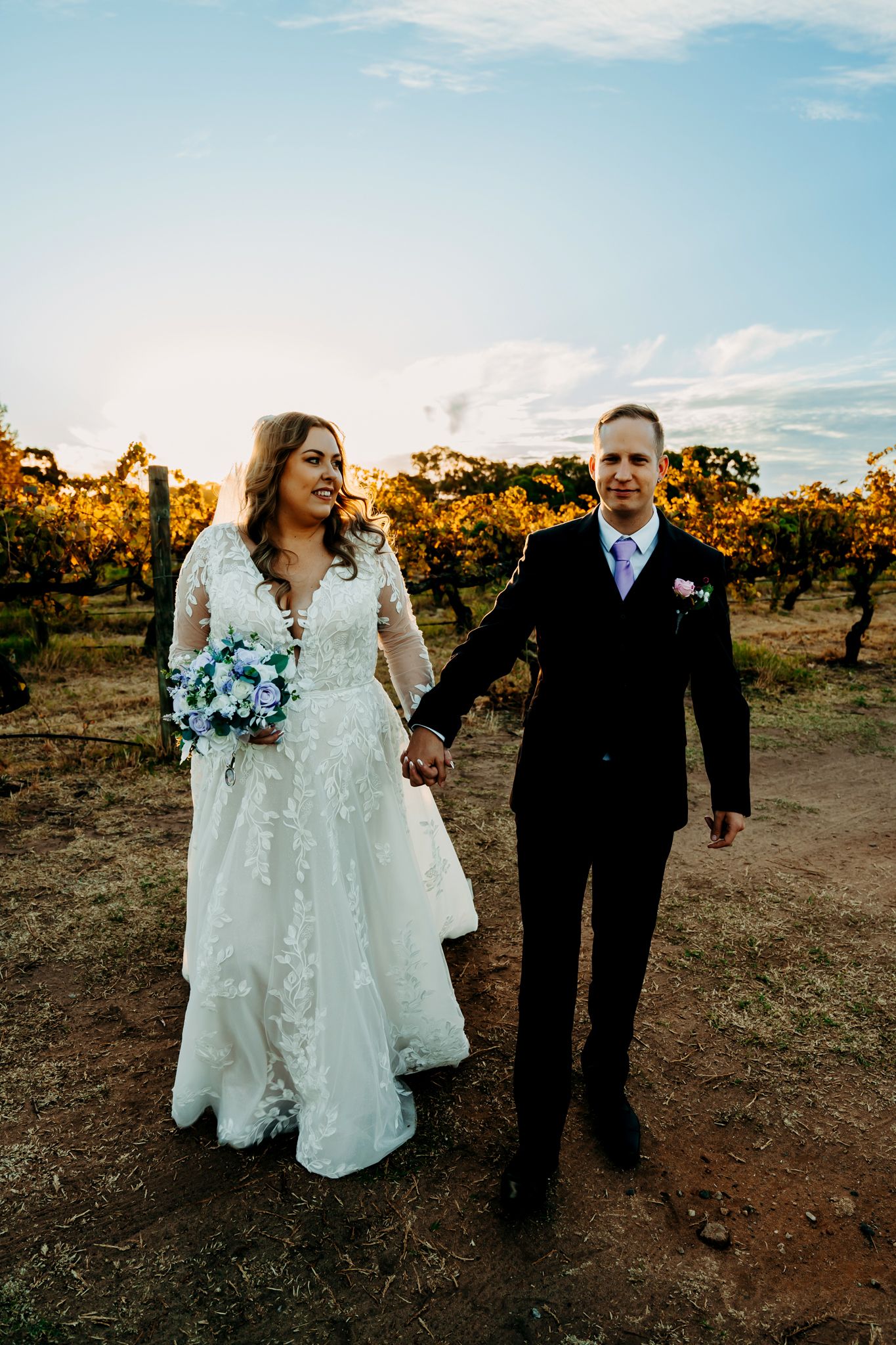 Newlyweds walking hand in hand through a rustic vineyard at sunset.
