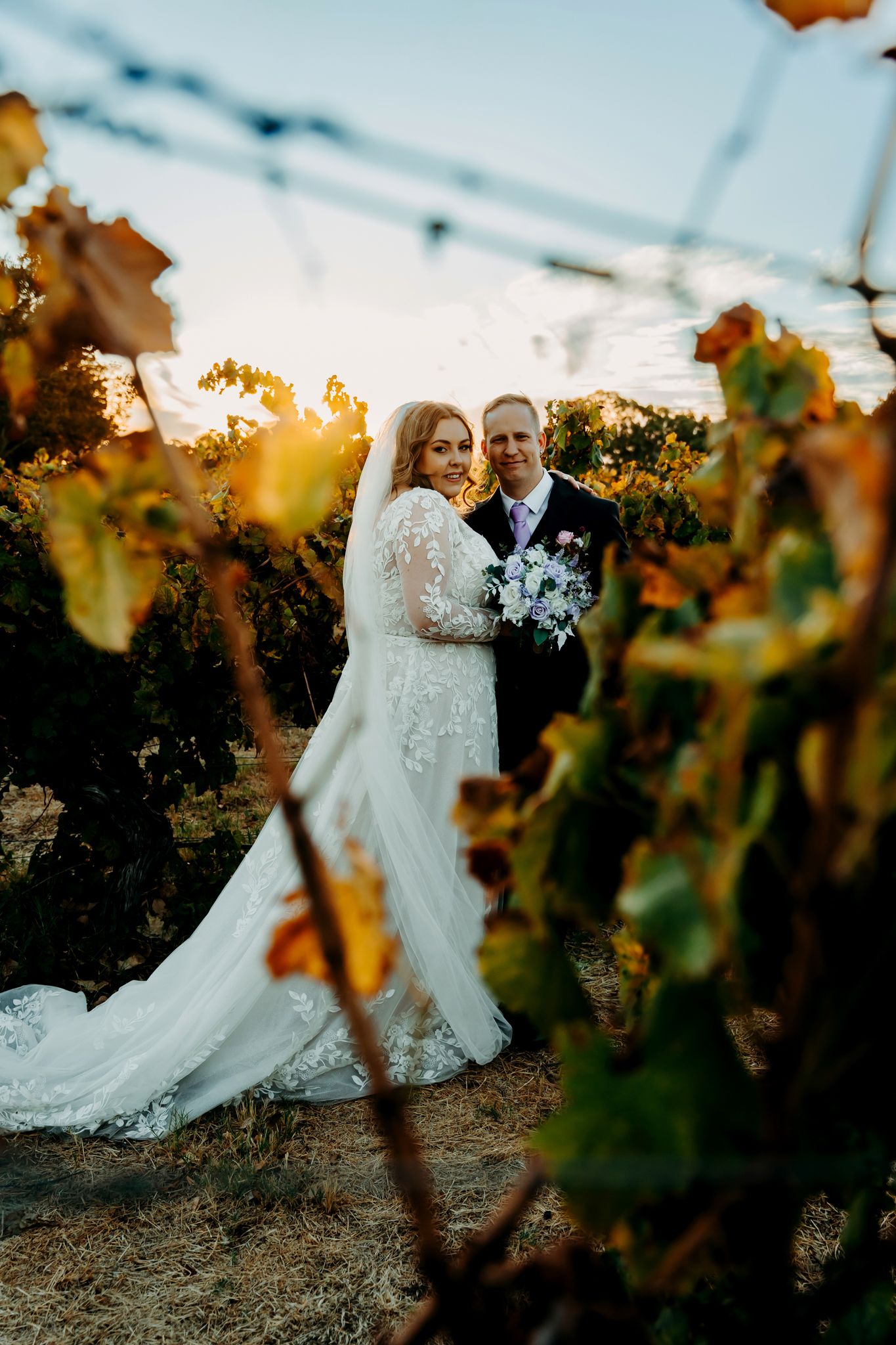 Elegant couple standing among vineyard vines at golden hour.