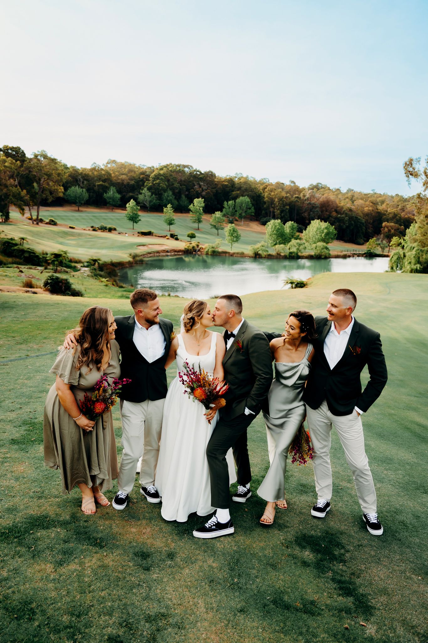 Wedding group shares candid moment on a lush golf course overlooking a scenic pond.