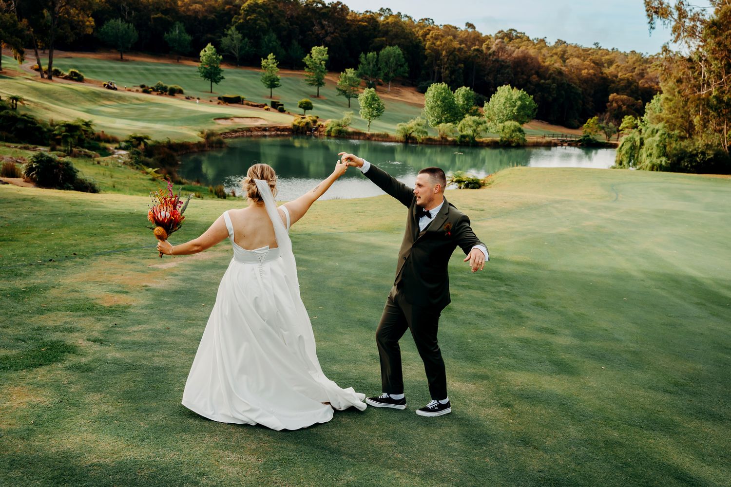 Newlyweds dance playfully on a golf course green with a scenic pond view.