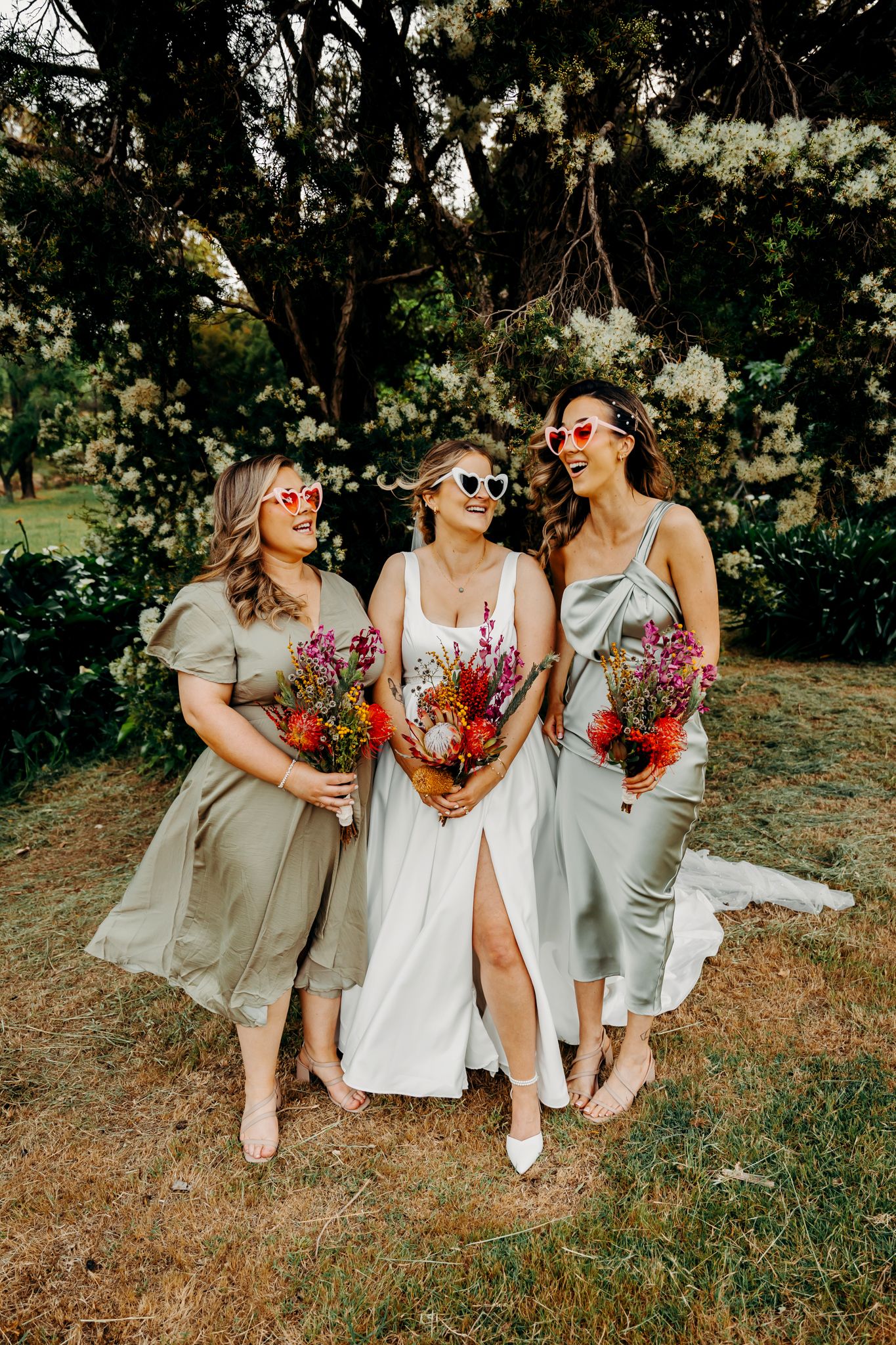 Three bridesmaids in flowing dresses share a laugh while holding vibrant bouquets in an outdoor garden setting.