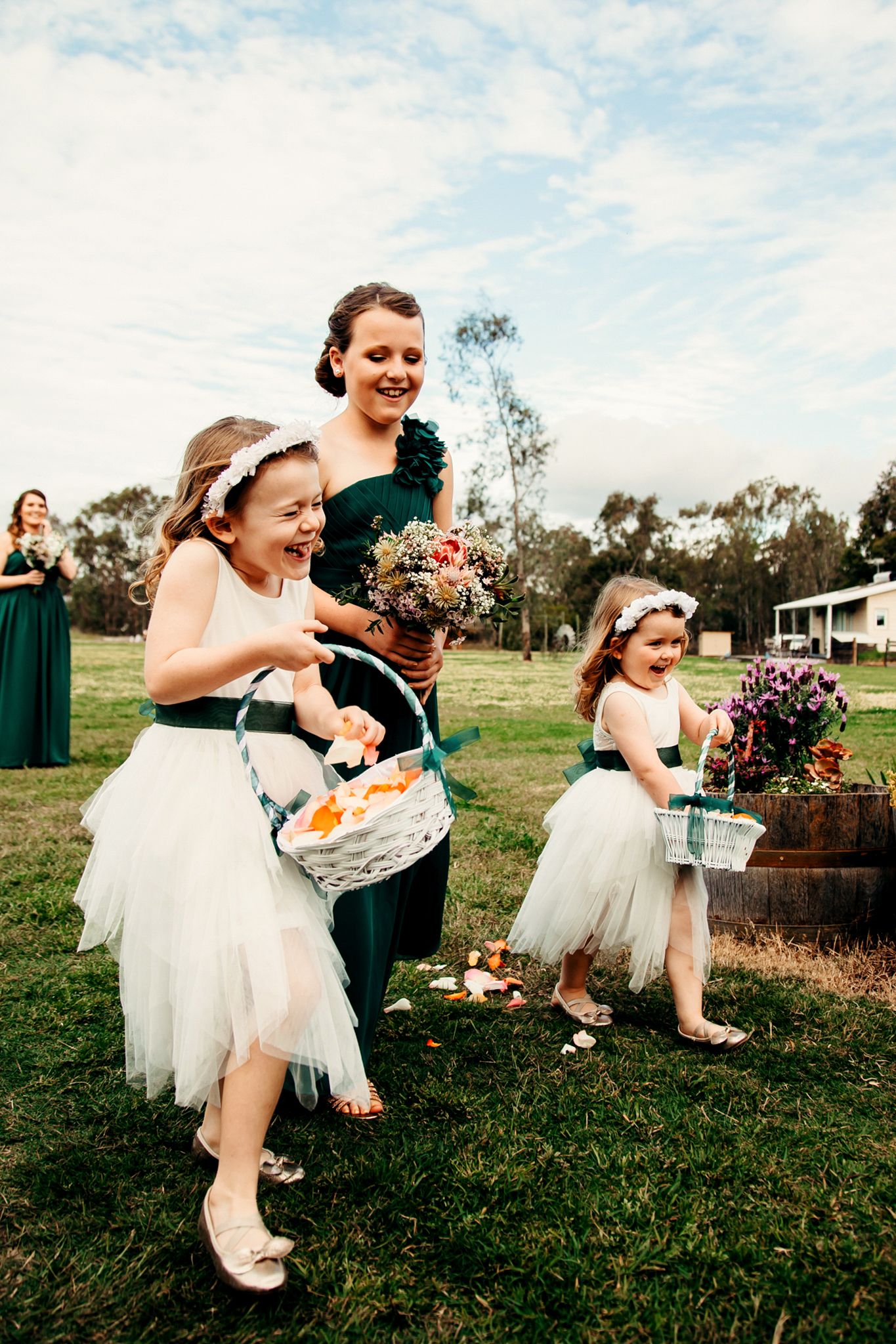 Flower girls in white tulle dresses carry baskets across field during outdoor wedding.