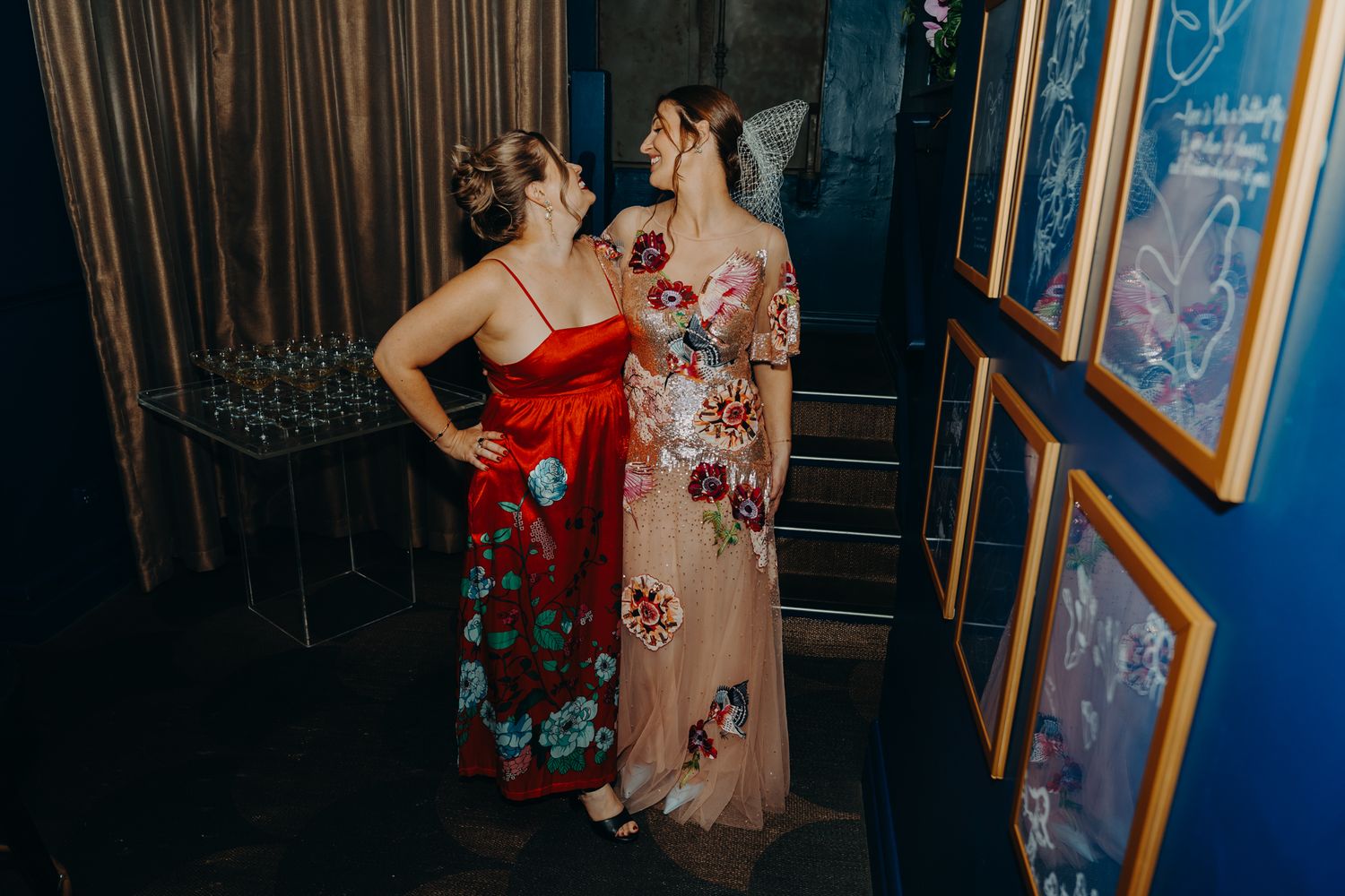 Two attendees in floral dresses stand near framed artwork at an event.