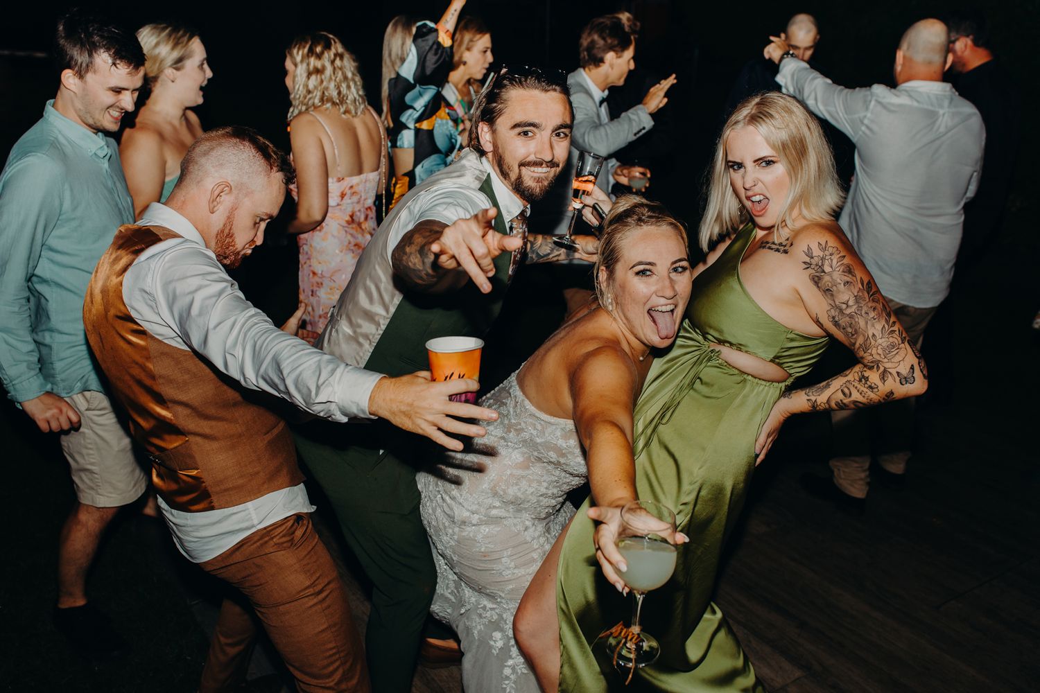 Wedding reception party guests dancing energetically on a dark dance floor with drinks in their hands.