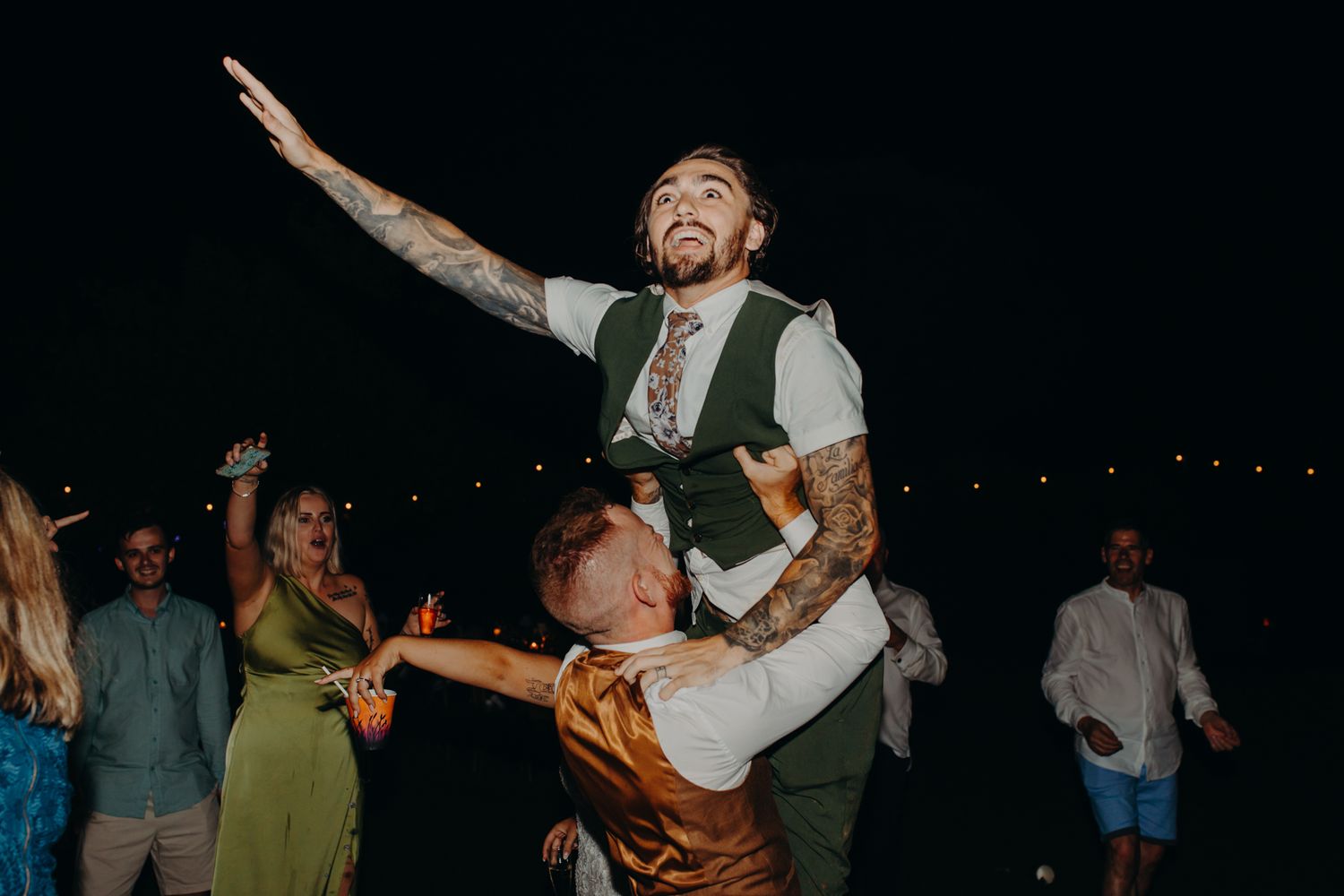 Wedding guest rides on shoulders during nighttime outdoor reception celebration on dance floor.